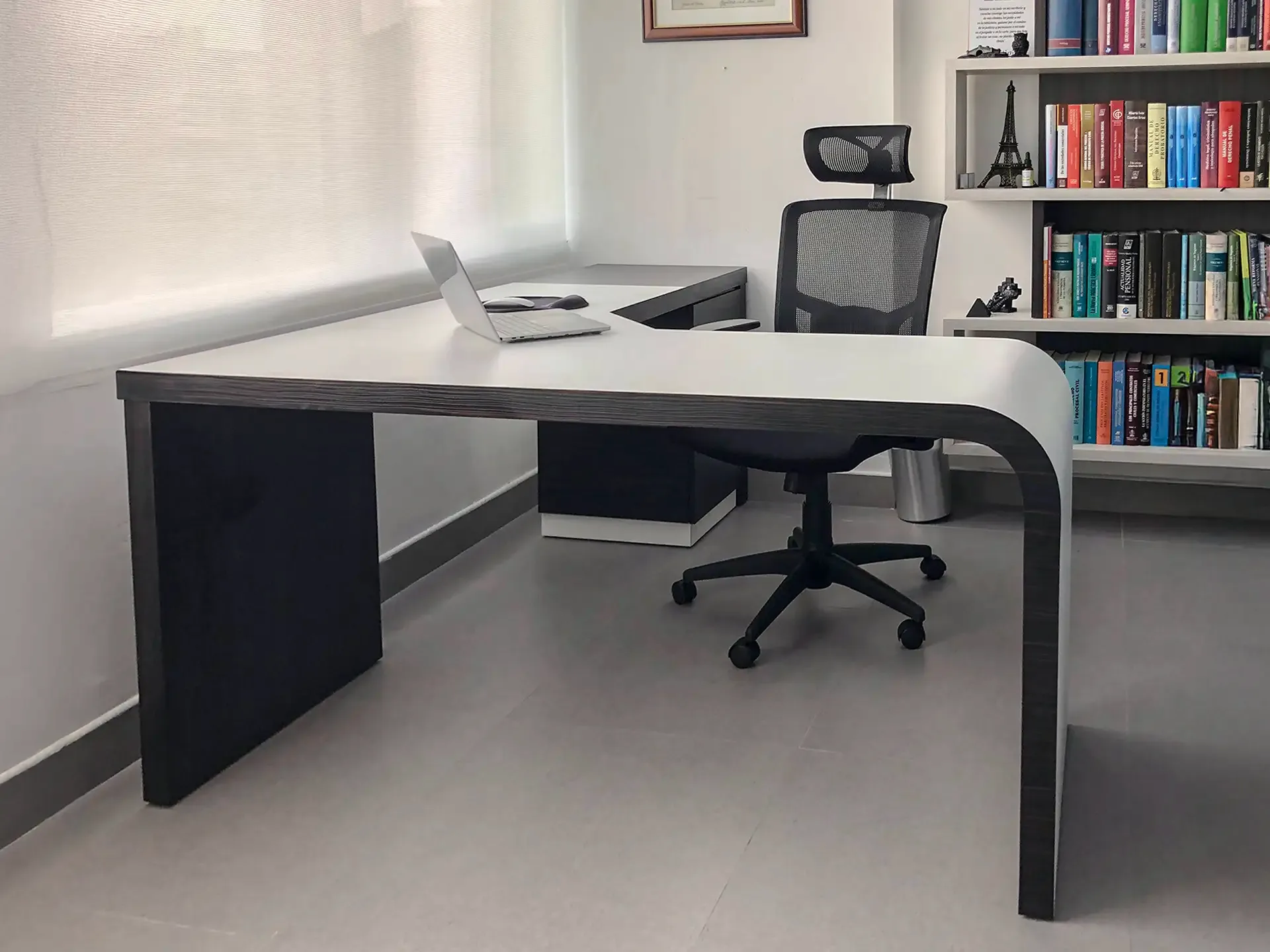 Escritorio blanco con superficie amplia, frente curvo oscuro, silla de oficina y biblioteca al fondo.